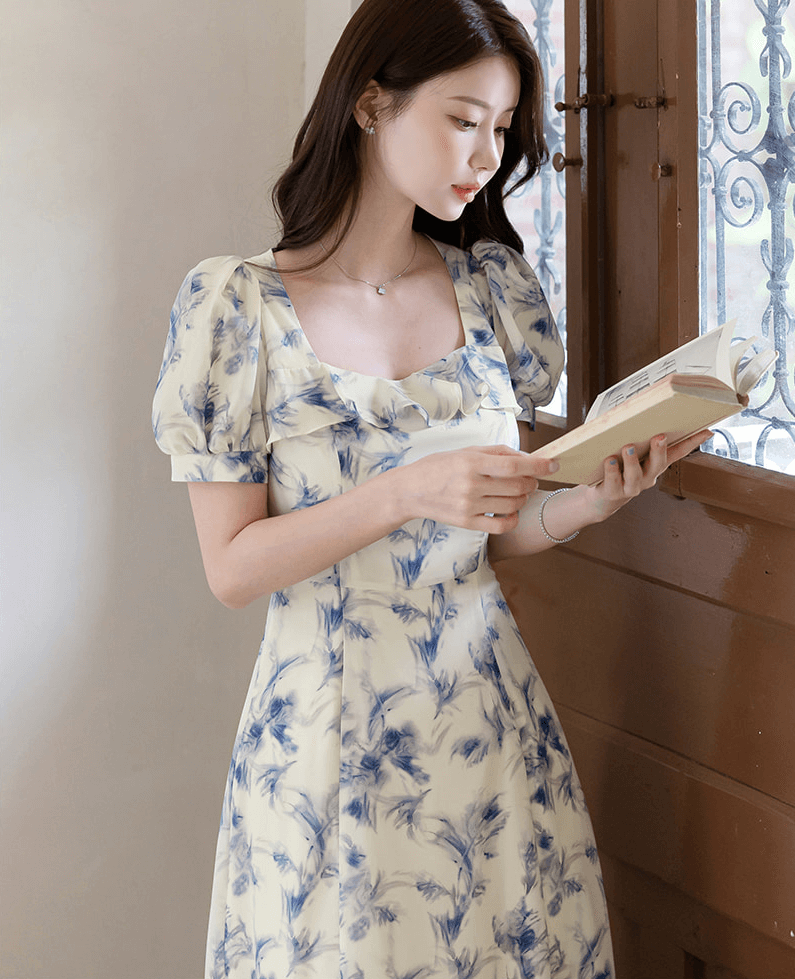Elegant chiffon dress with blue-violet abstract print and square ruffle neckline, worn by a woman reading by a window.