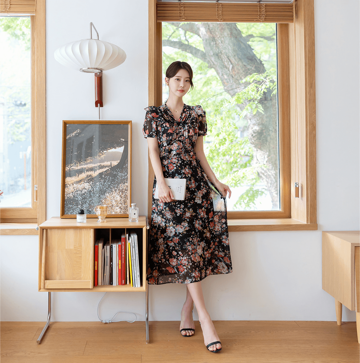 Woman wearing a floral chiffon dress with pintuck bodice and peplum detail, standing by a window in a stylish room.