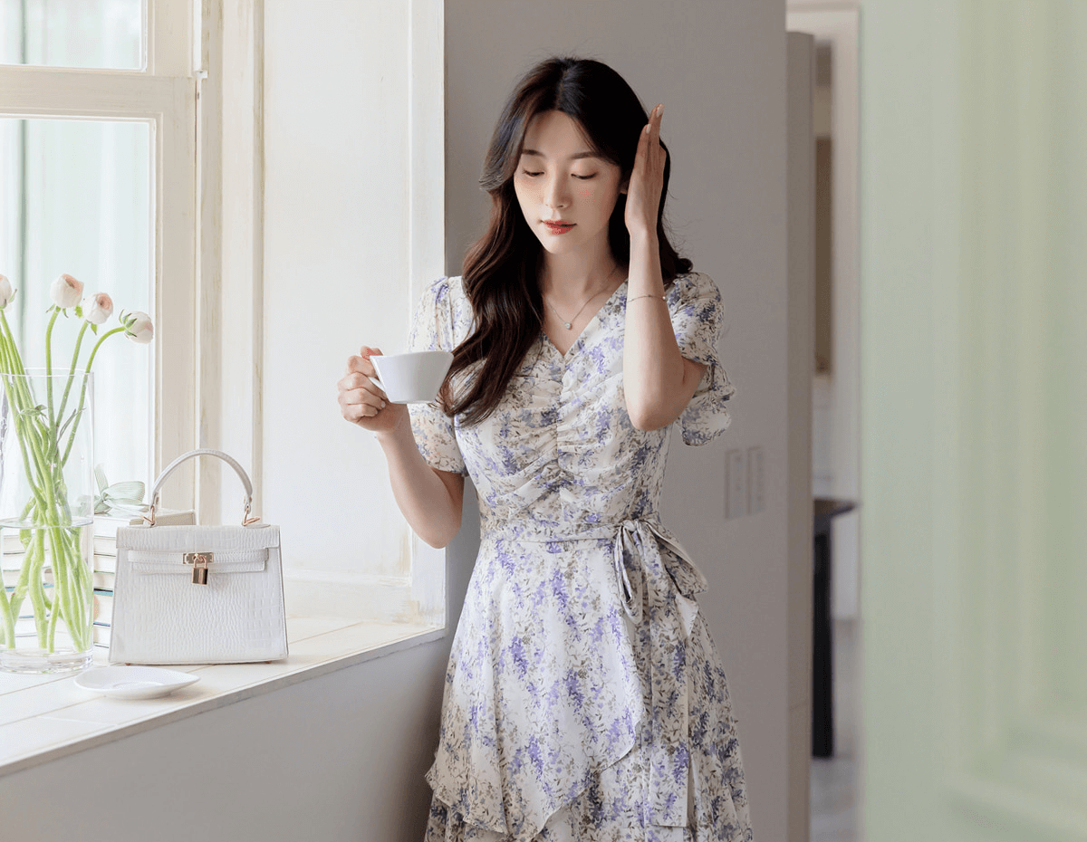Woman in asymmetrical tiered floral chiffon dress with purple blossoms, holding a coffee cup by a window, white handbag on the ledge.