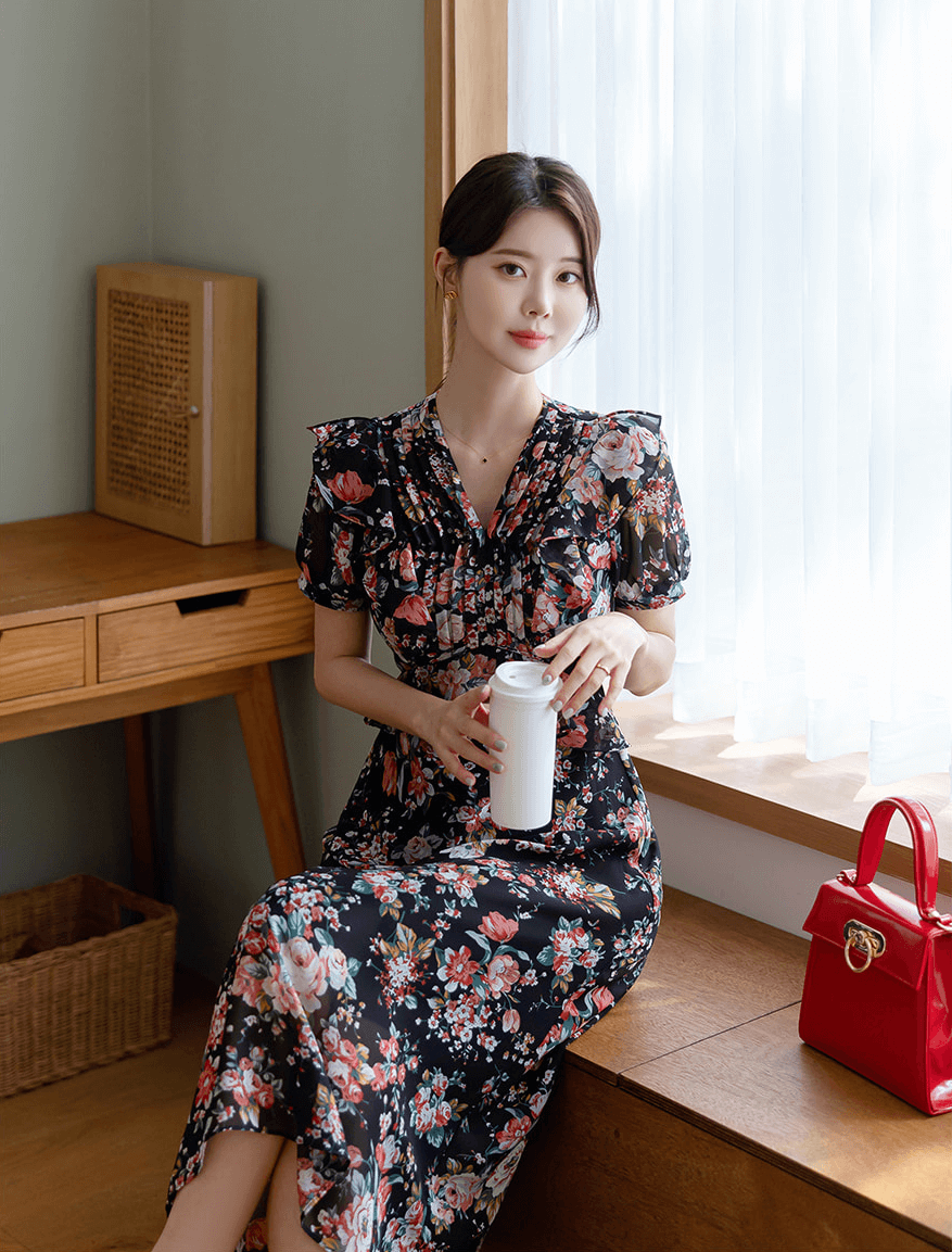 Woman wearing a floral chiffon dress with pintuck bodice and peplum detail, seated by a window, holding a tumbler, red purse nearby.