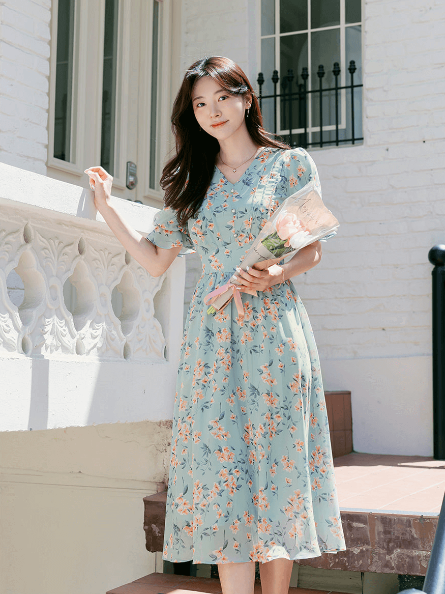 Woman wearing V-neck puff sleeve floral chiffon dress in breezy blue, standing outdoors holding flowers.