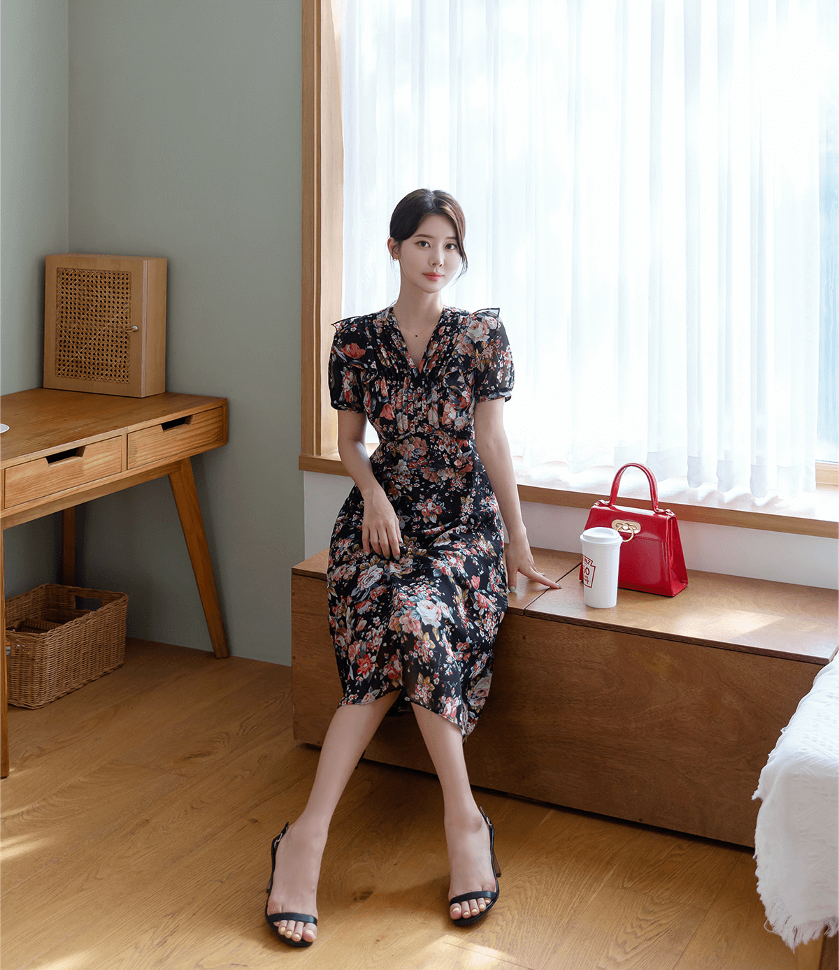 Woman wearing a floral chiffon dress with pintuck bodice and peplum detail, sitting by a window with a red handbag on a wooden bench.
