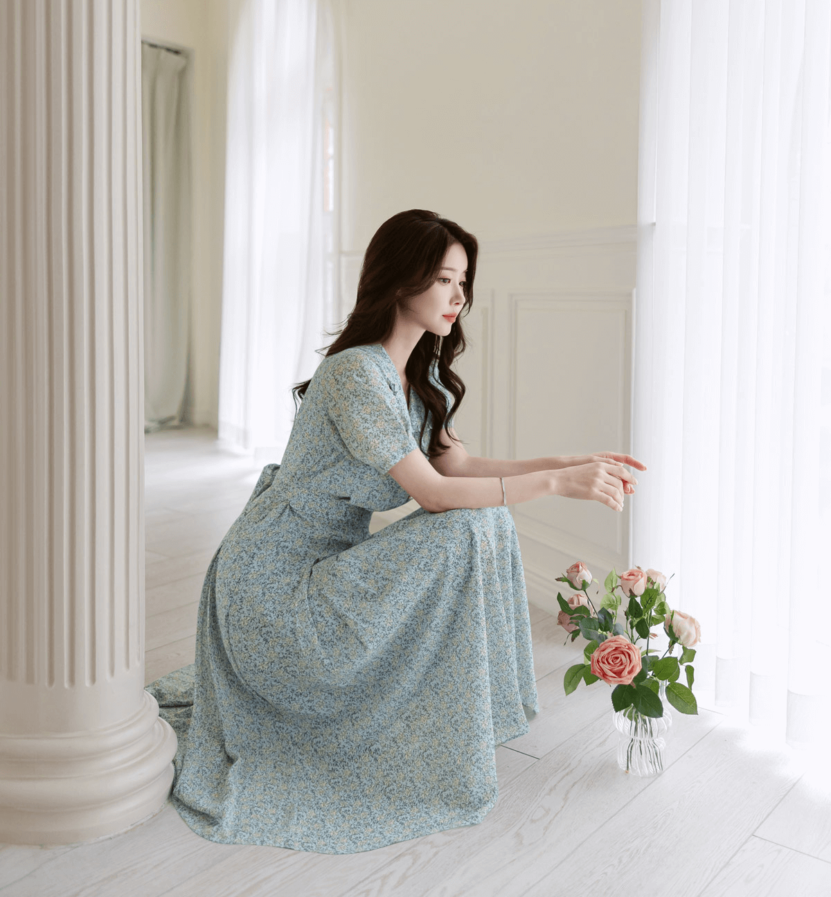 Woman in romantic floral chiffon dress with frill collar, sitting by a window with a vase of roses. Elegant fashion look.