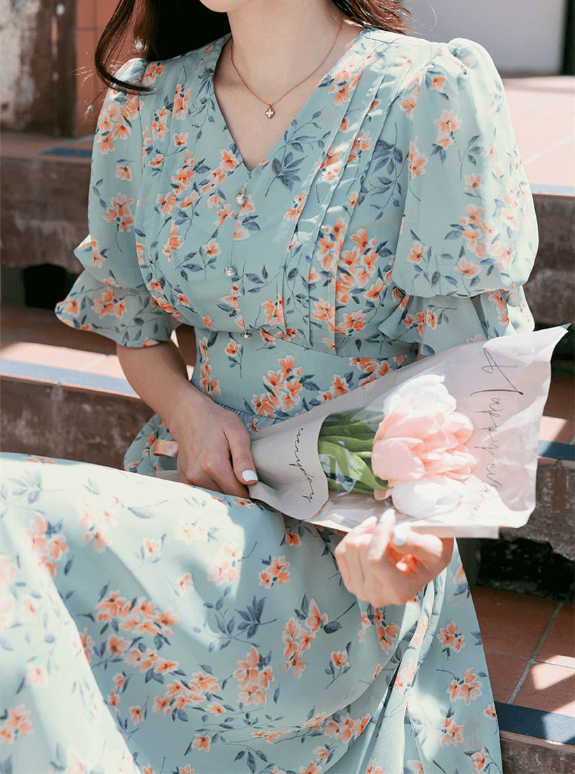 Woman in V-neck chiffon floral dress with puff sleeves sitting on steps holding pink flowers.