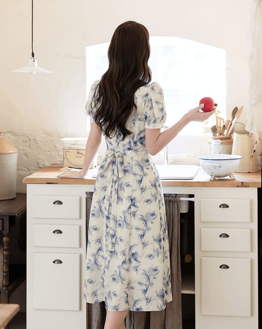 Woman in elegant blue-violet abstract print chiffon dress with ruffle neckline, standing in rustic kitchen with a red apple.