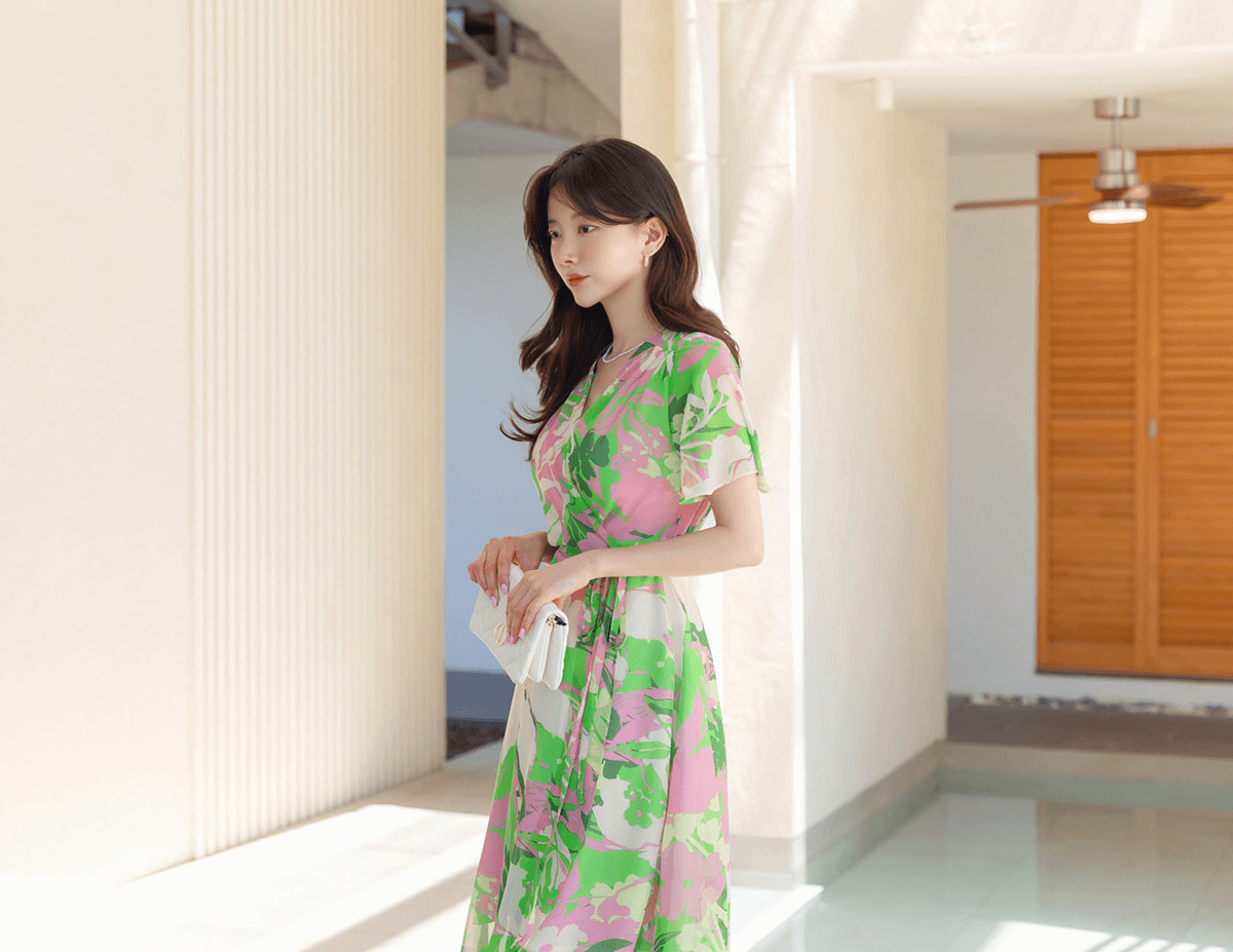 Woman wearing a chiffon wrap dress with tulip hem and floral print in green, yellow, and pink, standing in a sunny, modern hallway.