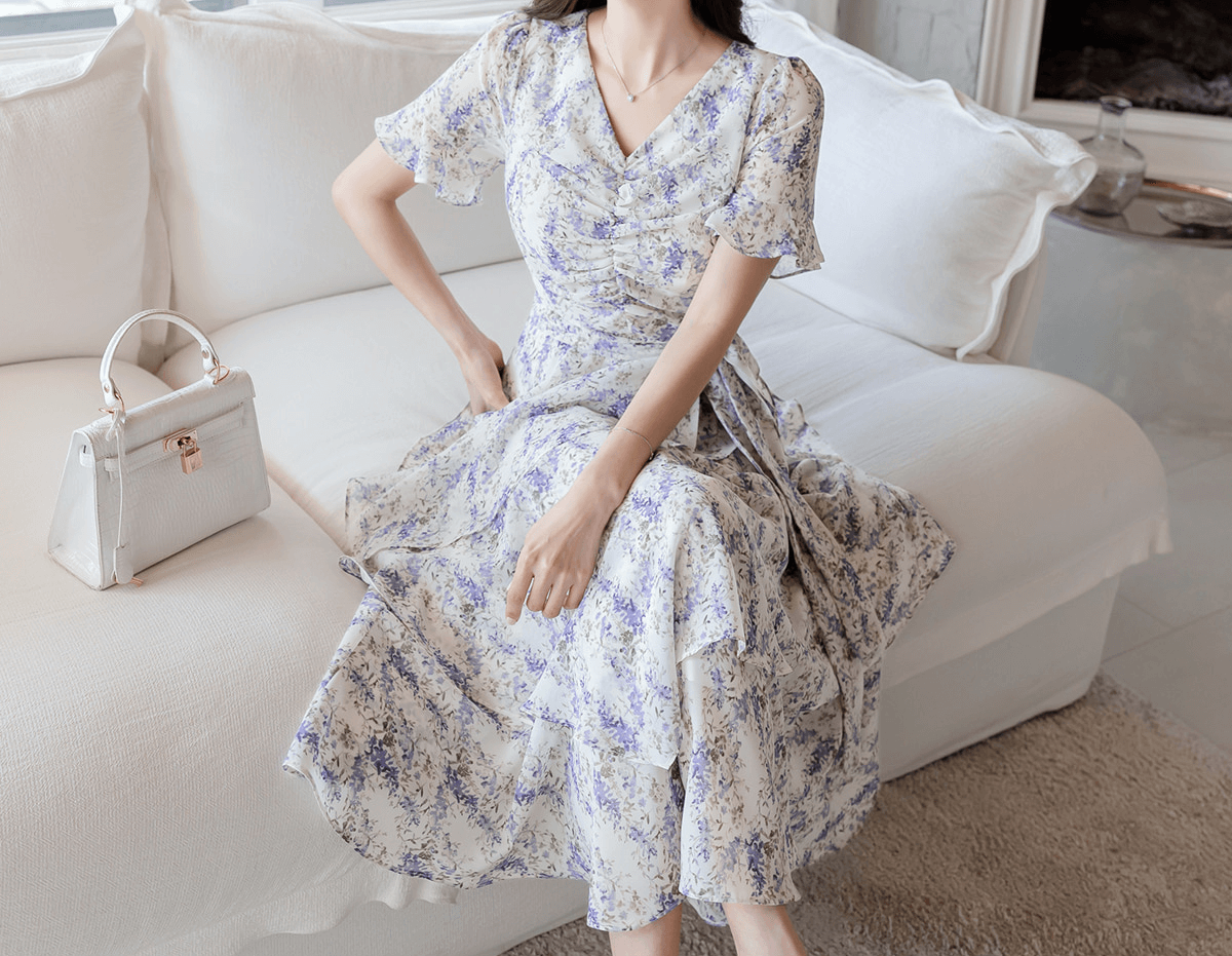 Woman wearing asymmetrical tiered floral chiffon dress with purple blossoms, seated on a white sofa next to a white handbag.