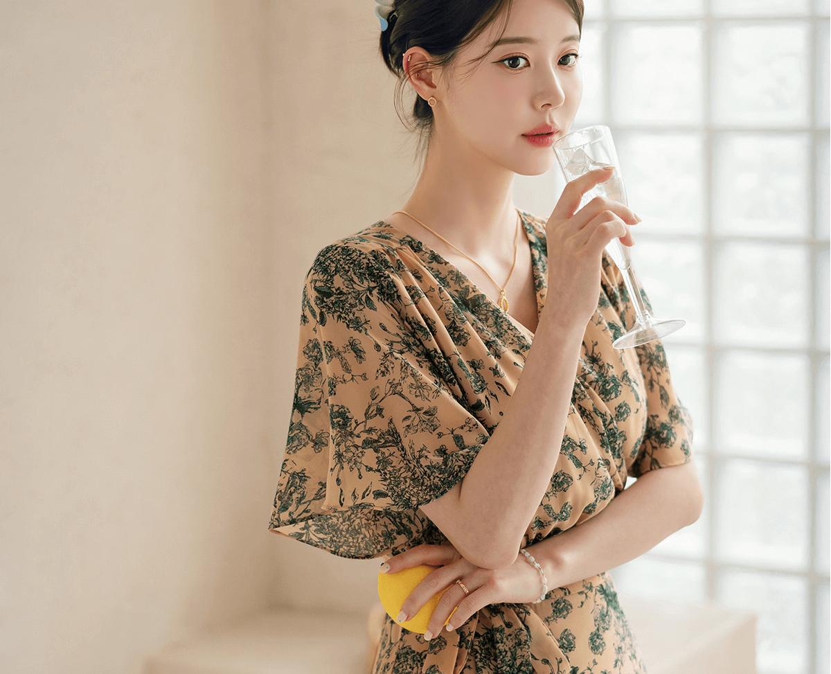 Woman wearing goddess-inspired wrap dress with wing sleeves, holding a champagne glass, near a sunlit window.