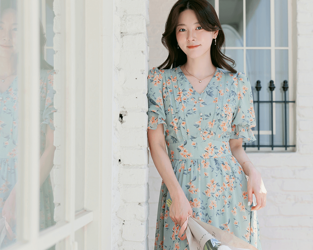 Woman wearing V-neck puff sleeve floral chiffon dress in breezy blue, standing near window with soft natural light and vibrant floral pattern.