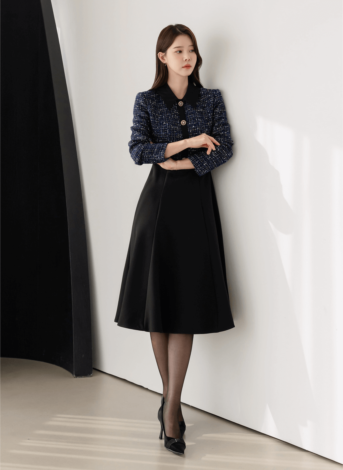 Woman wearing a two-tone tweed and black flare dress with gold button detail, standing against a white wall.