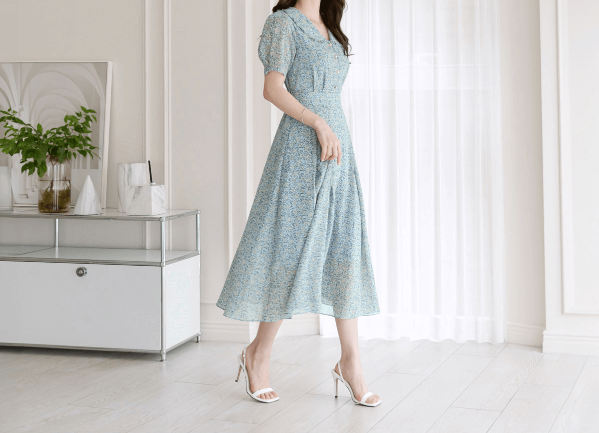 Woman wearing a romantic floral chiffon dress with frill collar and waist ribbon, standing in a bright room with white curtains.