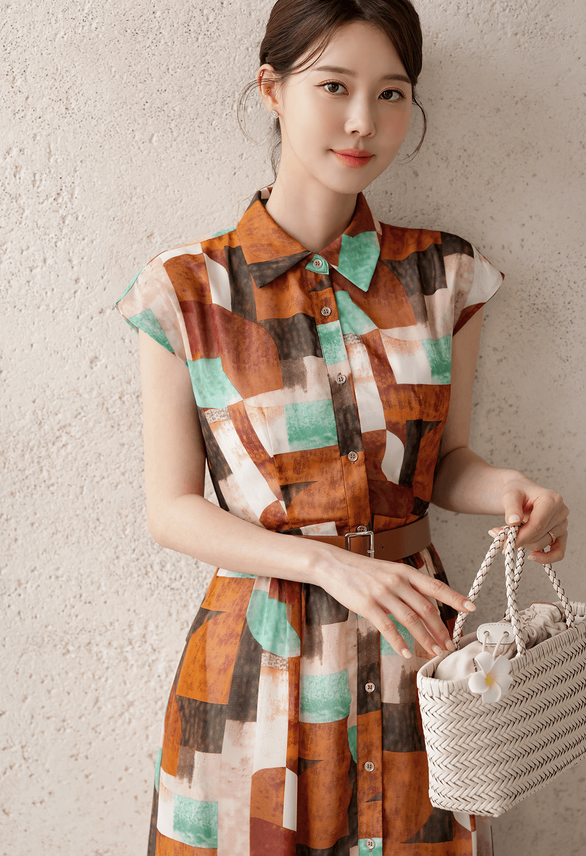 Woman wearing a patterned shirt dress with cap sleeves, classic collar, and a faux leather belt, holding a woven bag.