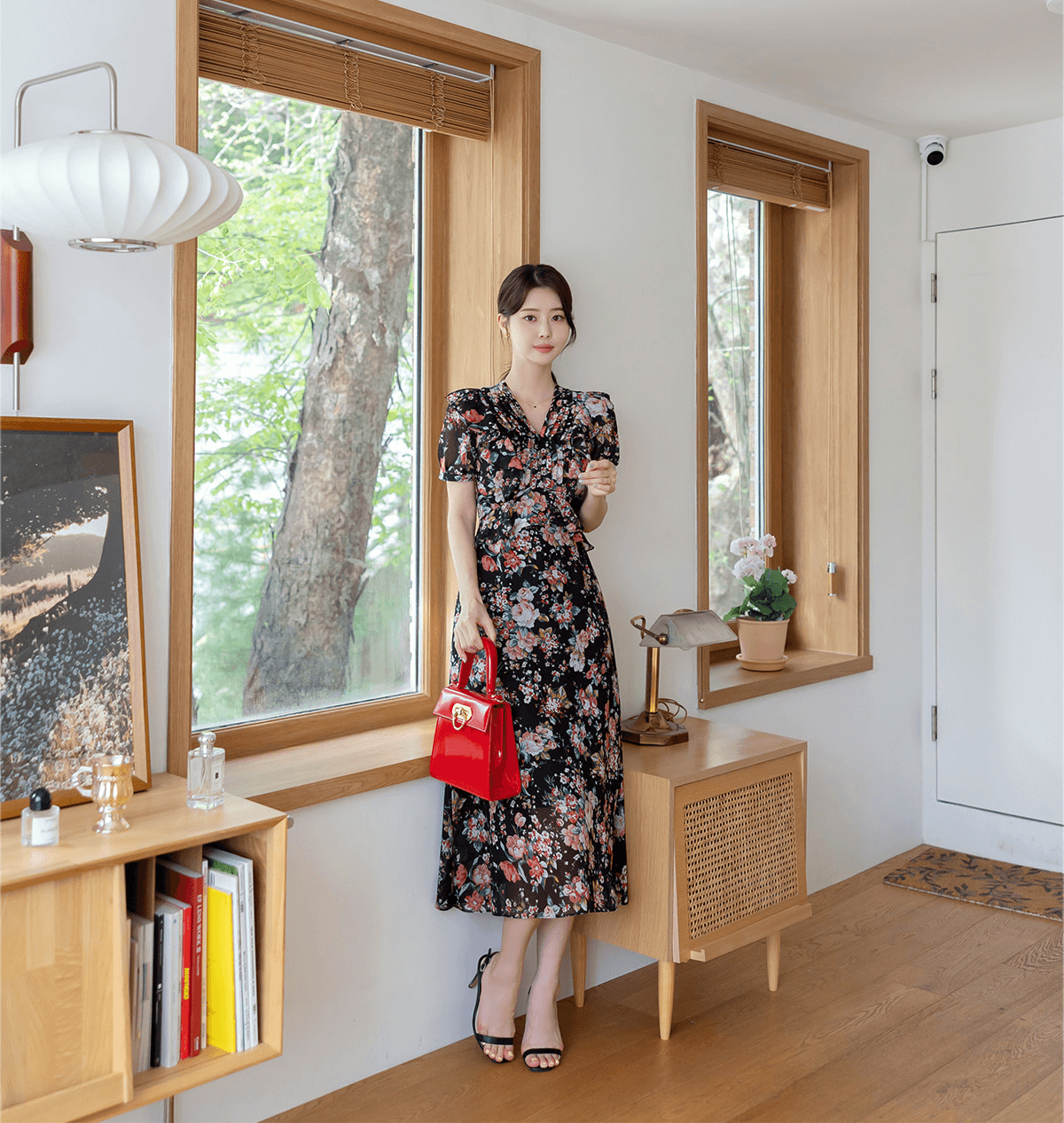 Woman wearing a floral chiffon dress with pintuck bodice and peplum detail, standing in a bright room with wooden accents.
