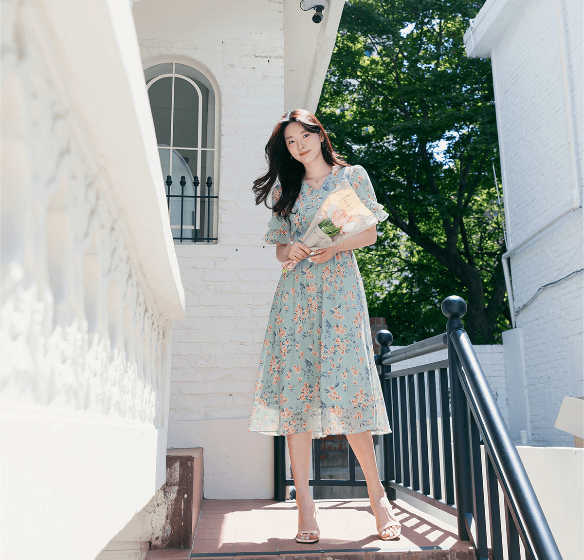 Woman wearing a V-neck puff sleeve floral chiffon dress in breezy blue, perfect for spring, standing outside a white house.
