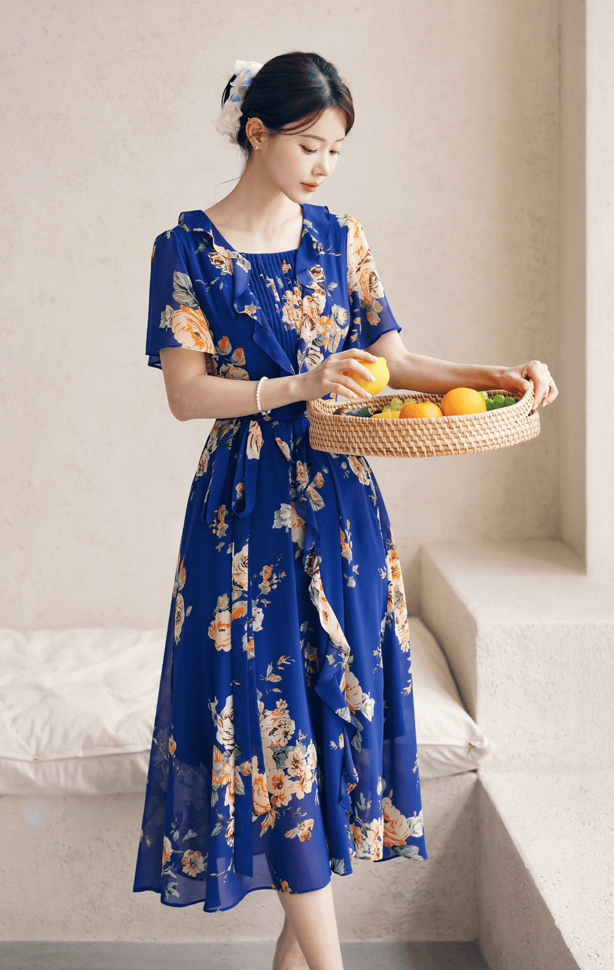 Woman in a blue floral chiffon dress with square neckline and sheer wing sleeves, holding a basket of fruit.
