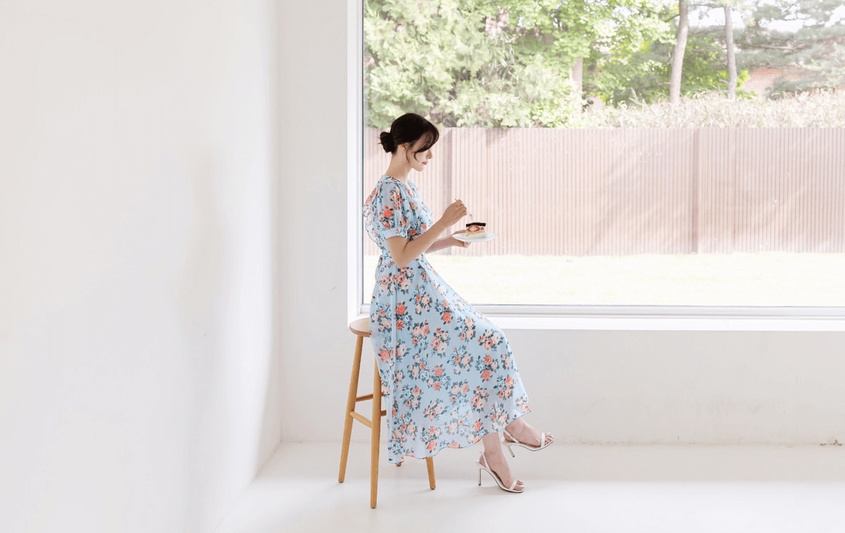 Woman wearing a romantic floral chiffon dress with short puff sleeves and V-neckline, sitting by a large window.