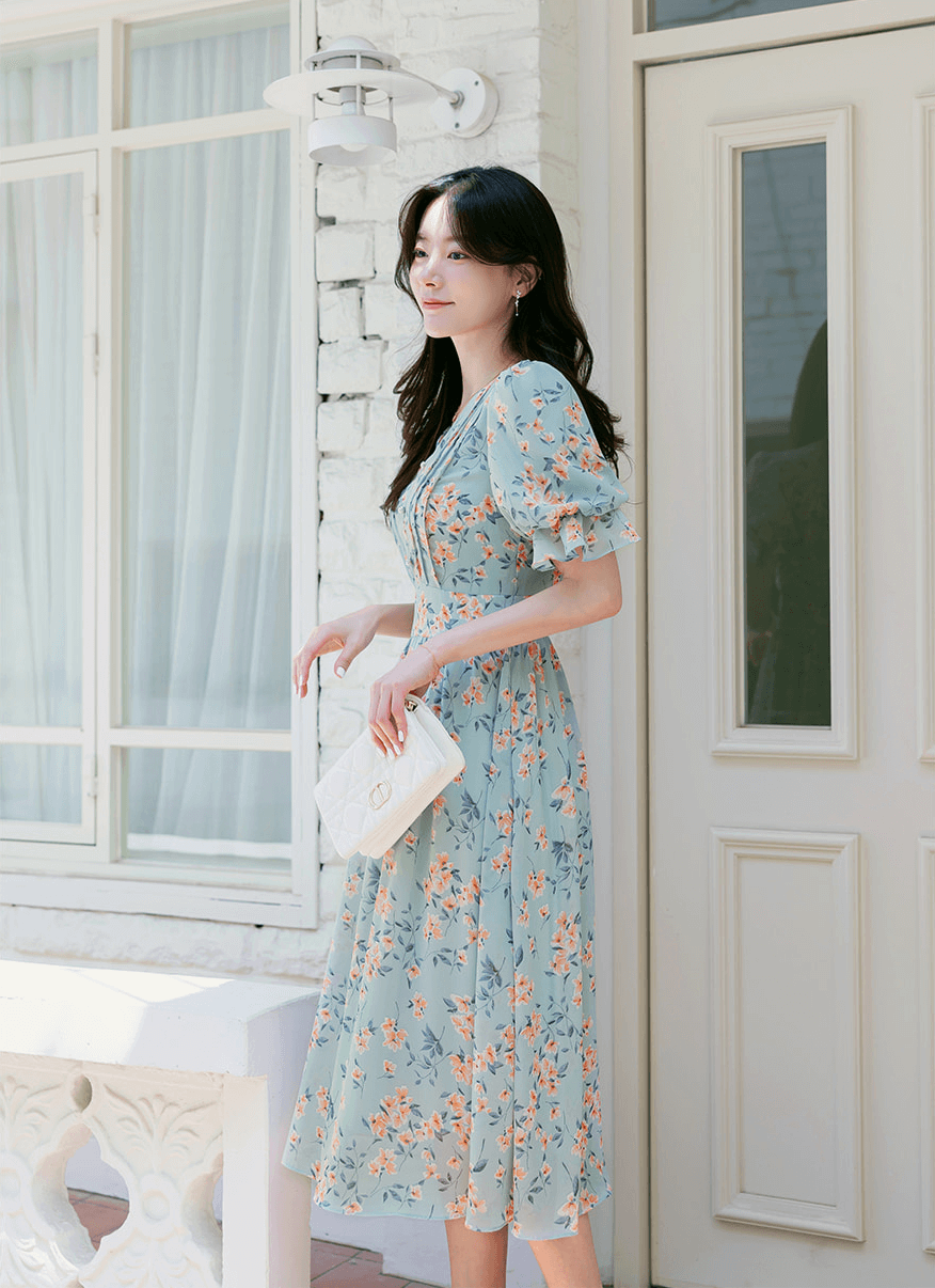 Woman wearing V-neck puff sleeve floral chiffon dress in breezy blue, standing outdoors with white clutch.