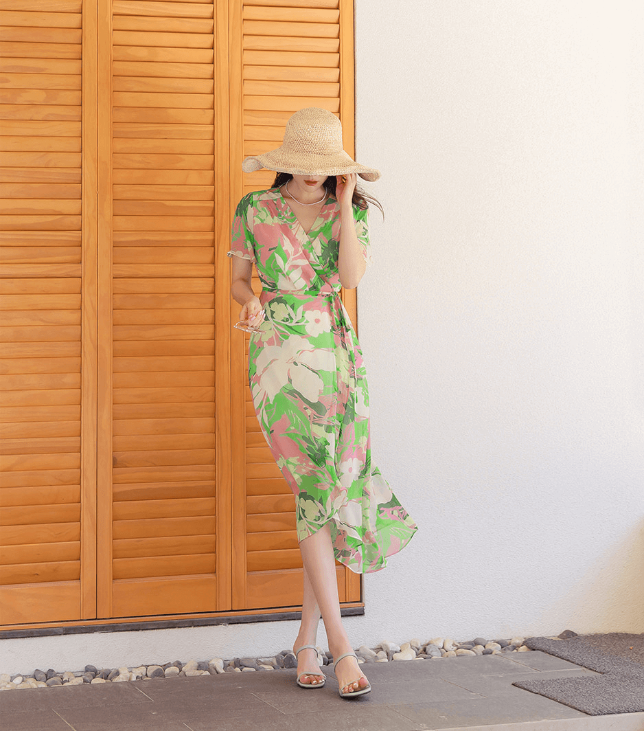Woman wearing a chiffon wrap dress with tulip hem and floral pattern, standing by wooden shutters, holding a hat.