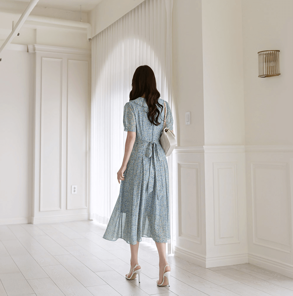 Woman wearing romantic floral chiffon dress with frill collar and waist ribbon, standing in a bright room.