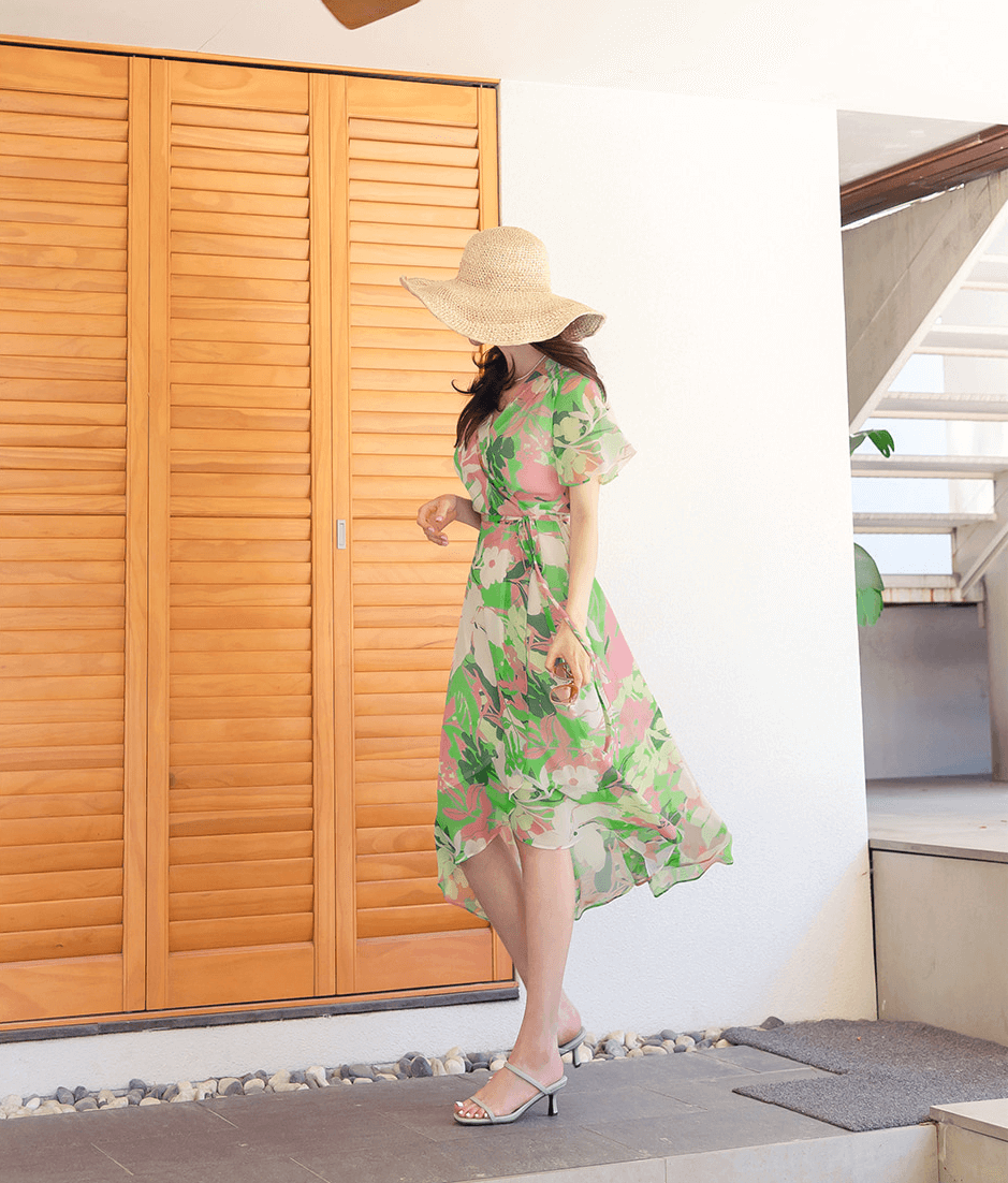 Woman in a green floral chiffon wrap dress with tulip hem, walking in a sunny outdoor setting.