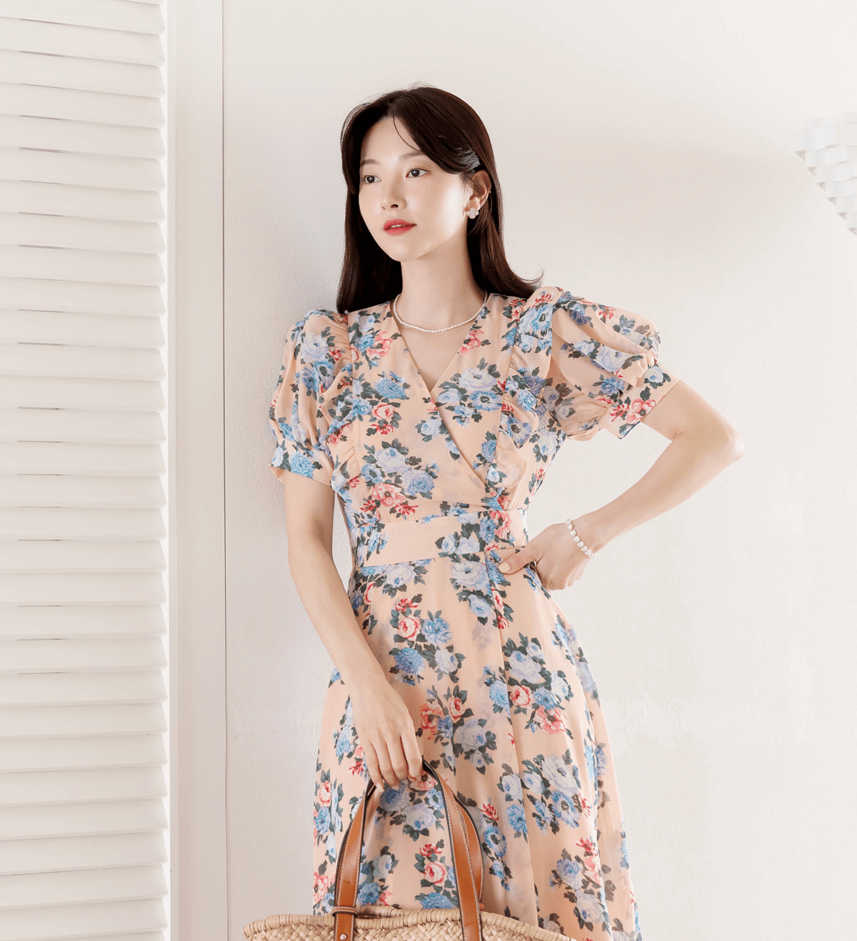 Woman wearing a romantic floral chiffon dress with puff sleeves and V-neckline, holding a bag, against light background.