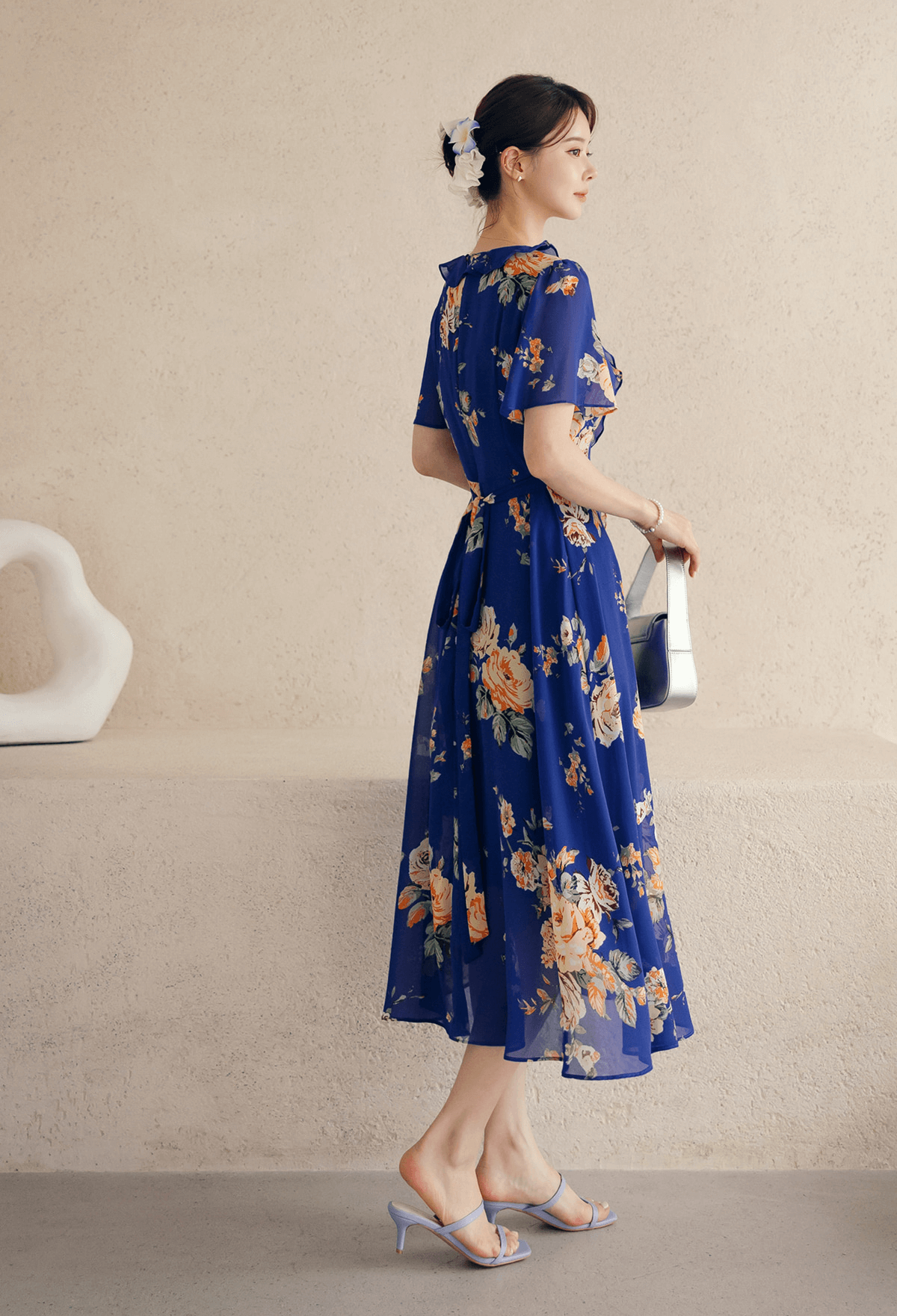 Woman wearing a blue floral chiffon dress with sheer wing sleeves and square neckline, standing sideways holding a clutch.