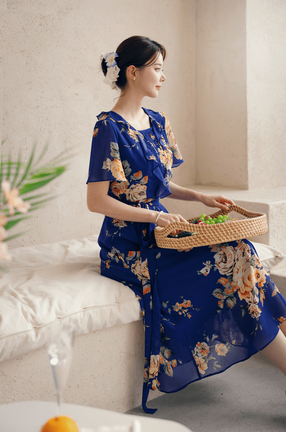 Woman wearing a floral chiffon dress with sheer wing sleeves and square neckline, sitting with a fruit basket.