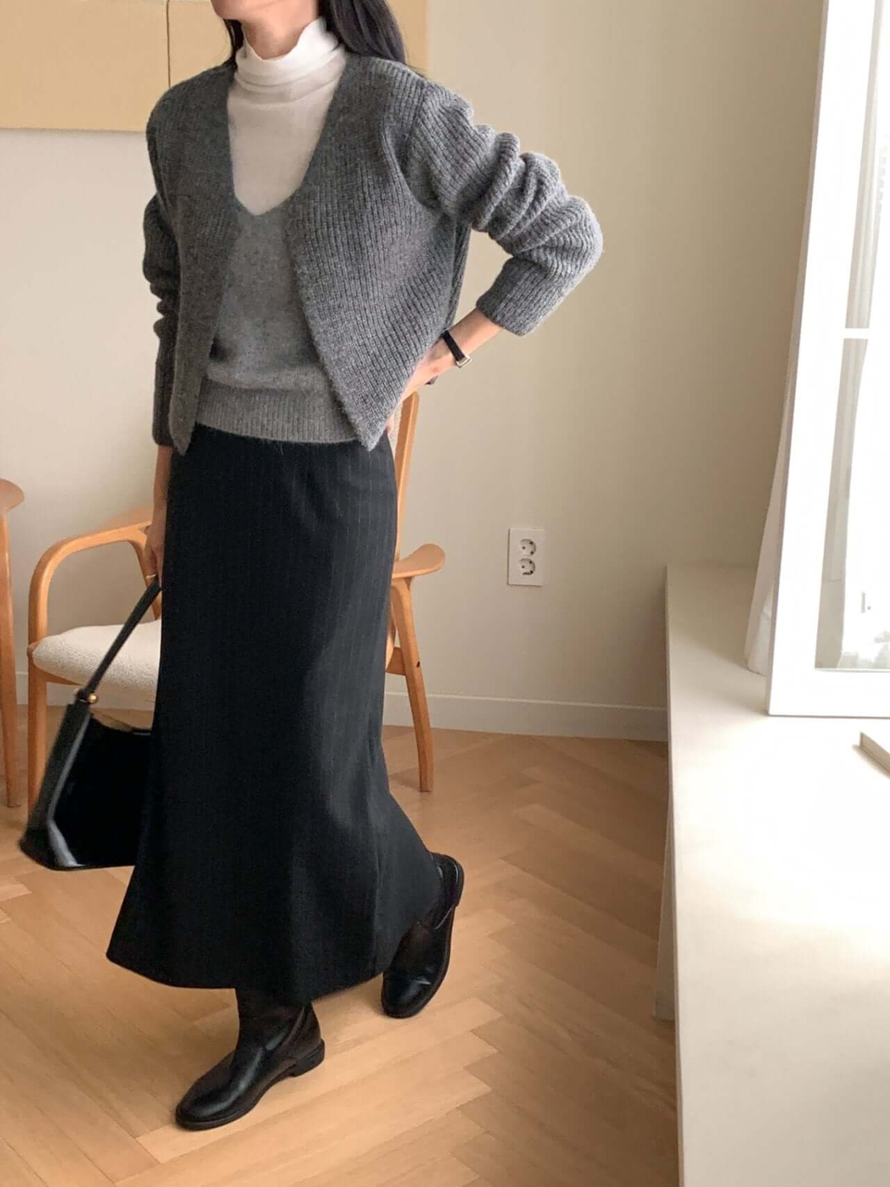 Woman wearing a gray alpaca blend cardigan, styled with a black skirt and white turtleneck in a cozy interior.