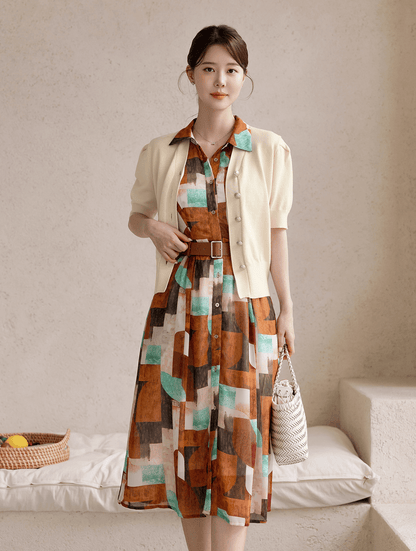 Woman wearing a patterned shirt dress with cap sleeves, classic collar, and faux leather belt, standing in a minimalist setting.