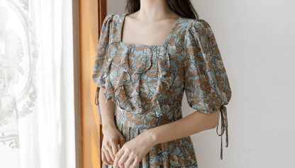 Woman wearing a pleated dress with floral pattern, square neckline, and ribbon-tied slit sleeves, standing by a window.