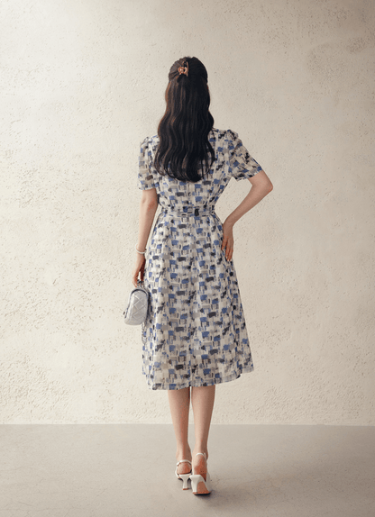 Back view of woman wearing breezy chiffon shirt dress with abstract blue pattern and belt, holding a small purse, in a light room