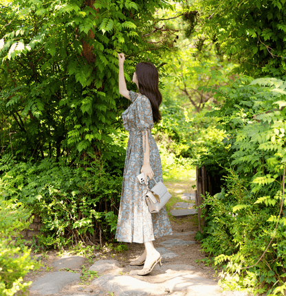 Woman in floral pleated dress with slit sleeves and ribbon ties, walking in a vibrant green garden, holding a stylish handbag.