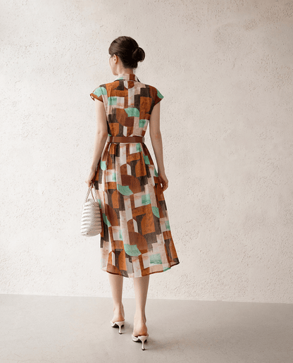 Back view of a woman wearing a patterned shirt dress with cap sleeves, classic collar, and faux leather belt, holding a woven bag.