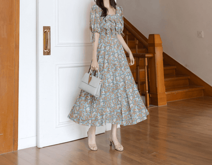 Woman wearing pleated dress with slit sleeves and ribbon ties, standing beside staircase, holding a white handbag.